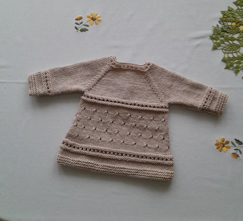 May include: A beige knitted baby dress with a delicate lace pattern. The dress has long sleeves and a round neckline.