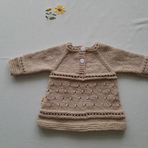 May include: A beige knitted baby dress with long sleeves and a button closure. The dress has a delicate lace pattern and a slightly flared skirt.