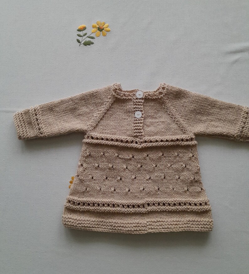 May include: A beige knitted baby dress with long sleeves and a button closure. The dress has a delicate lace pattern and a slightly flared skirt.