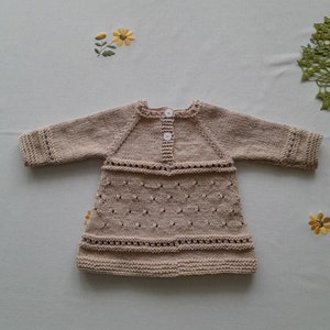 May include: A beige knitted baby dress with long sleeves and a button closure at the back.
