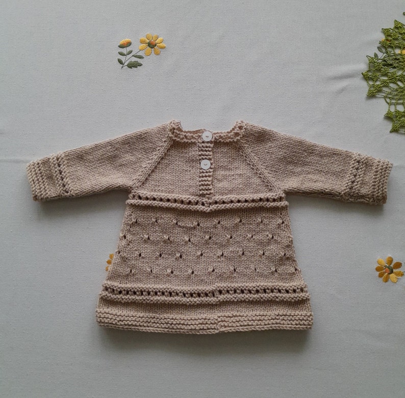 May include: A beige knitted baby dress with long sleeves and a button closure at the back.