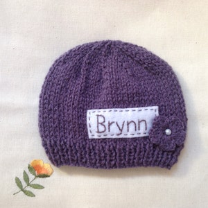 K&ouml;nnte beinhalten: Eine gestrickte lila Baby-M&uuml;tze mit dem Namen "Brynn" auf einem wei&szlig;en Aufn&auml;her gestickt. Eine kleine lila Blume mit Perlenmitte ist an der Seite befestigt. Die M&uuml;tze liegt auf einer cremefarbenen Oberfl&auml;che.