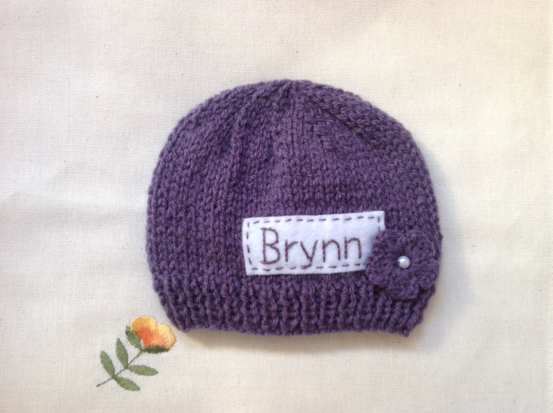 K&ouml;nnte beinhalten: Eine gestrickte lila Baby-M&uuml;tze mit dem Namen "Brynn" auf einem wei&szlig;en Aufn&auml;her gestickt. Eine kleine lila Blume mit Perlenmitte ist an der Seite befestigt. Die M&uuml;tze liegt auf einer cremefarbenen Oberfl&auml;che.