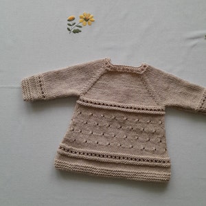 May include: A beige knitted baby dress with long sleeves and a delicate lace pattern.