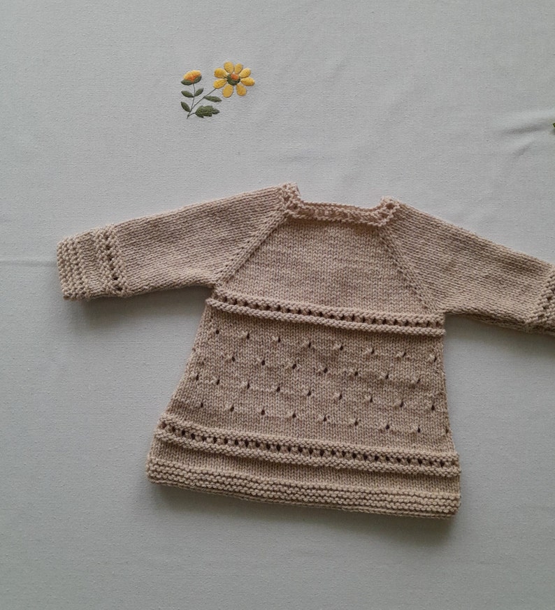 May include: A beige knitted baby dress with long sleeves and a delicate lace pattern.