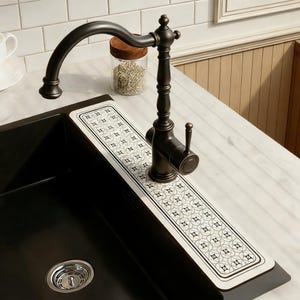 May include: A dark bronze kitchen faucet with a curved spout and a decorative handle. A rectangular sink protector with a white and black geometric pattern sits beneath the faucet. A black sink is visible.