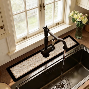 May include: A kitchen sink with a black faucet and a decorative sink mat. The mat has a cream-colored floral pattern with gold accents and a black border. A window is visible in the background.