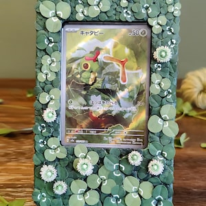 Puede incluir: Un marco de fotos decorativo con una carta de Pokémon en su interior. El marco está adornado con adornos verdes en forma de trébol y pequeños acentos de flores blancas. La carta presenta un personaje verde con texto japonés.