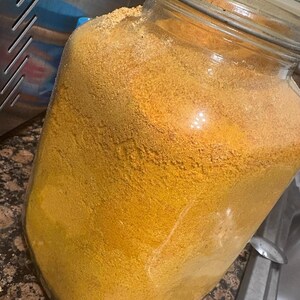 May include: A large, clear glass jar filled with a fine, yellow-orange powder. The jar has a metal clasp and a glass lid. The powder appears to be a spice or seasoning, possibly turmeric or curry powder, and fills the jar to the top.