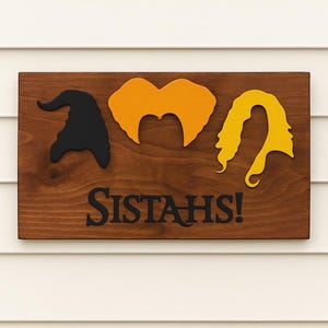 Puede incluir: Letrero de madera con la palabra "SISTAH'S!" y siluetas de un sombrero de bruja, pelo naranja y pelo amarillo. El letrero est&aacute; montado sobre un fondo de revestimiento blanco. Decoraci&oacute;n de Halloween.