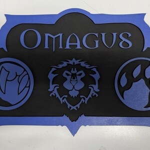 May include: A black and blue wooden sign with the name "OMAGUS" in the center. The sign features three circular emblems: a claw, a lion's head, and a set of claws. The sign has a blue border.