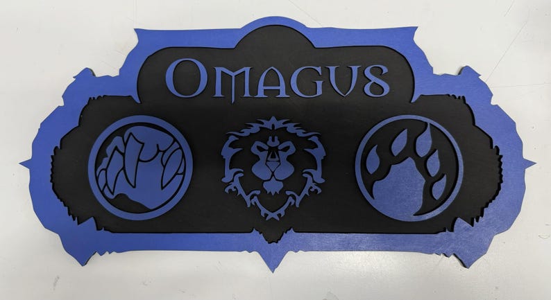 May include: A black and blue wooden sign with the name "OMAGUS" in the center. The sign features three circular emblems: a claw, a lion's head, and a set of claws. The sign has a blue border.