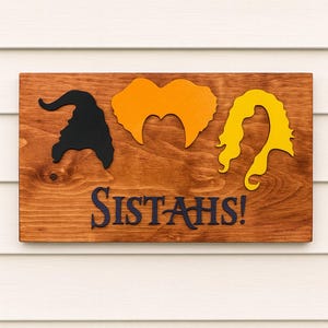 Puede incluir: Letrero de madera con siluetas de tres peinados de brujas en negro, naranja y amarillo. El texto azul oscuro dice "SISTAH'S!". El letrero est&aacute; montado sobre un fondo de revestimiento blanco.