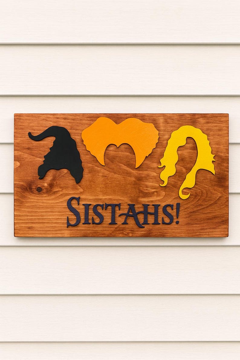 Puede incluir: Letrero de madera con siluetas de tres peinados de brujas en negro, naranja y amarillo. El texto azul oscuro dice "SISTAH'S!". El letrero est&aacute; montado sobre un fondo de revestimiento blanco.