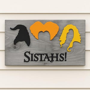 Puede incluir: Letrero de madera con fondo gris que presenta siluetas negras de un sombrero de bruja, cabello naranja y cabello amarillo. La palabra "SISTAH'S!" est&aacute; escrita en letras negras debajo de las siluetas. El letrero est&aacute; montado sobre un revestimiento blanco.