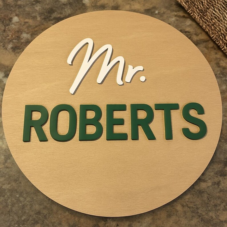 May include: A round wooden sign with the text "Mr. ROBERTS". The word "Mr." is in a white script font, and "ROBERTS" is in a bold, green font. The sign has a light brown wood grain background.