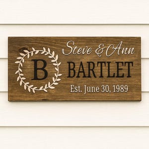 Puede incluir: Letrero de madera rectangular con los nombres "Steve & Ann BARTLET" y la fecha "Est. June 30, 1989". El letrero tiene un fondo marr&oacute;n oscuro con letras blancas y una corona decorativa alrededor de la letra "B".