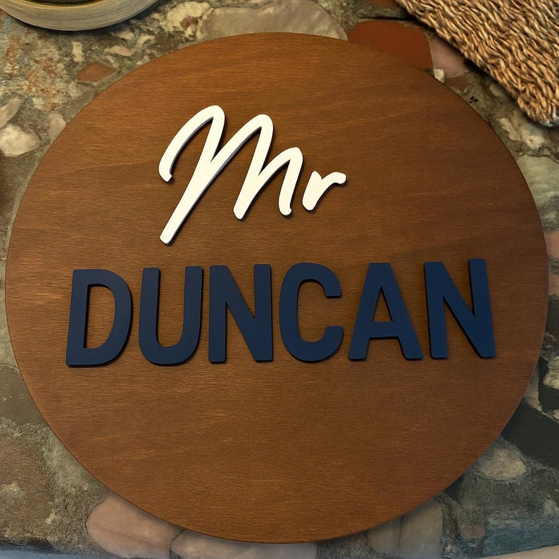 May include: A round wooden sign with the word "Mr" in white script above the name "DUNCAN" in navy blue block letters. The sign has a dark brown wood grain finish.