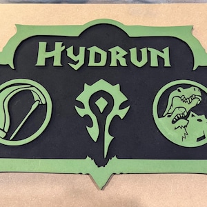 May include: A two-tone wooden sign with the name "HYDRUN" in green lettering. The sign features three green symbols on a black background, including a scythe, a stylized emblem, and a wolf-like creature.
