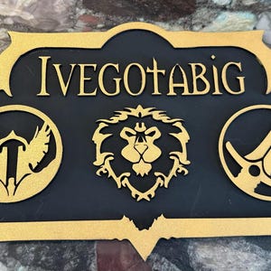 May include: A wooden sign with the text "IVEGOTABIG" in gold lettering. The sign has a black background and features three gold-colored emblems: a bird, a lion's head, and a crossed sword and shield.