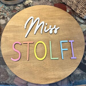 May include: A round wooden sign with the name "Miss STOLFI" in white and pastel colors. The sign has a light brown wood grain background and is likely for a teacher or classroom.