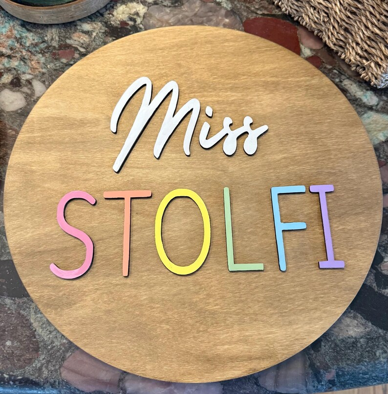 May include: A round wooden sign with the name "Miss STOLFI" in white and pastel colors. The sign has a light brown wood grain background and is likely for a teacher or classroom.