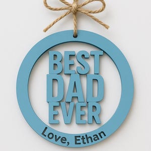 May include: A round, light blue wooden sign with the words "BEST DAD EVER" in bold, raised letters. The sign includes the inscription "Love, Ethan" and is suspended by a natural twine bow.