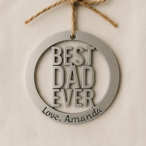 May include: A silver metal ornament with the words "BEST DAD EVER" in bold letters. The words "Love, Amanda" are inscribed below. The ornament is suspended by a twine bow.