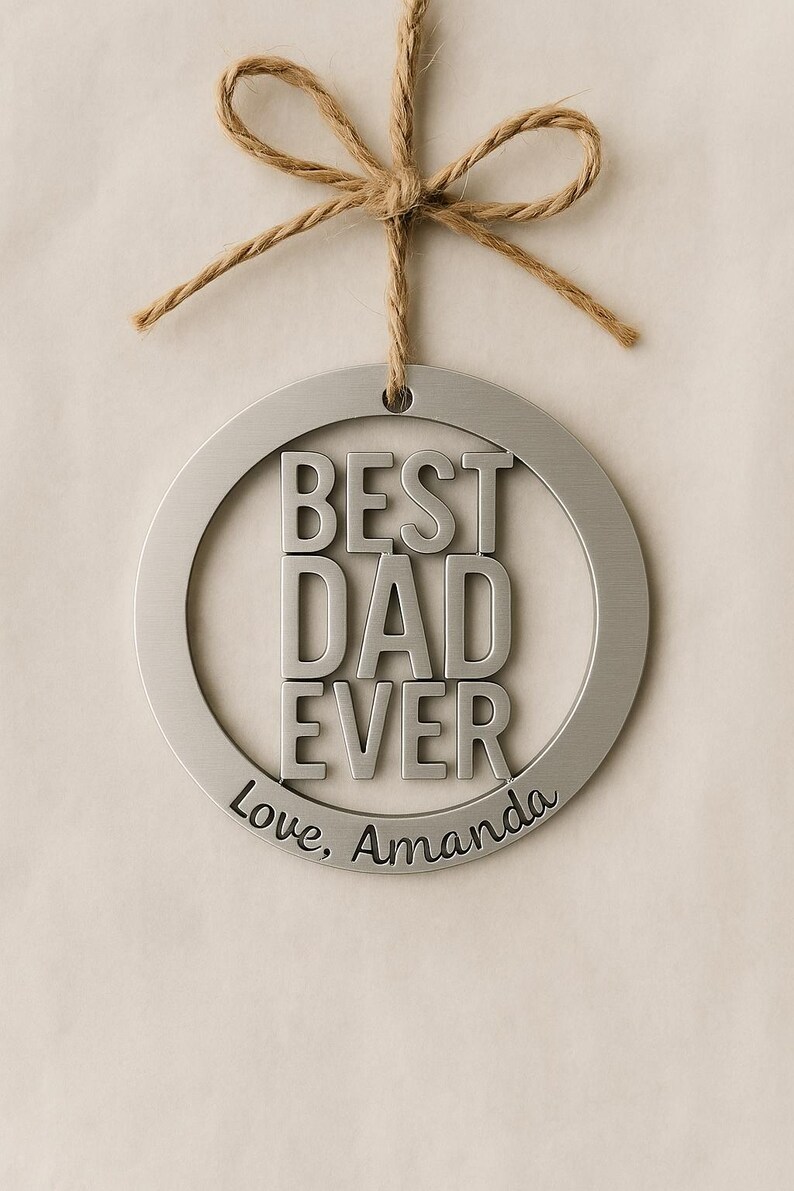 May include: A silver metal ornament with the words "BEST DAD EVER" in bold letters. The words "Love, Amanda" are inscribed below. The ornament is suspended by a twine bow.