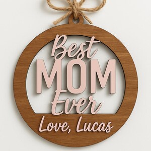 May include: Wooden round ornament with the words "Best MOM Ever Love, Lucas" in pink and brown. The ornament has a natural wood grain finish and is suspended by a twine loop.