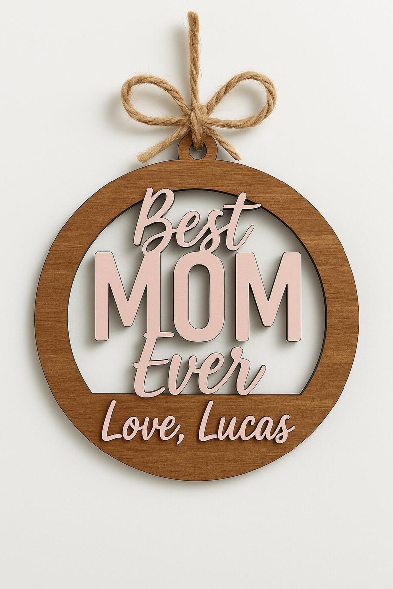 May include: Wooden round ornament with the words "Best MOM Ever Love, Lucas" in pink and brown. The ornament has a natural wood grain finish and is suspended by a twine loop.