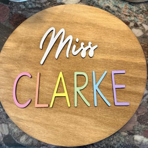 May include: A round wooden sign with the name "Miss Clarke" in colorful, raised letters. The word "Miss" is in white script, while "Clarke" is in pastel rainbow colors. The sign has a natural wood grain finish.
