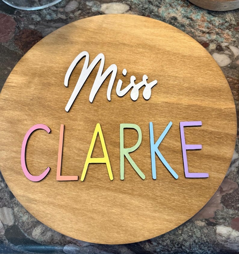 May include: A round wooden sign with the name "Miss Clarke" in colorful, raised letters. The word "Miss" is in white script, while "Clarke" is in pastel rainbow colors. The sign has a natural wood grain finish.
