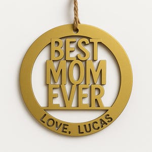 May include: A gold-colored circular ornament with the words "BEST MOM EVER" in bold letters. The text "LOVE, LUCAS" is inscribed at the bottom. A twine loop is attached at the top for hanging.
