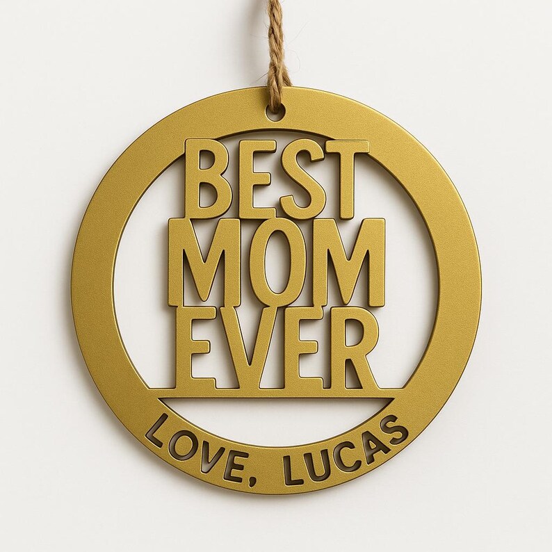 May include: A gold-colored circular ornament with the words "BEST MOM EVER" in bold letters. The text "LOVE, LUCAS" is inscribed at the bottom. A twine loop is attached at the top for hanging.