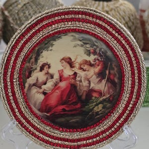 May include: A decorative plate with a painted scene of women in red and white dresses. The plate has a red and gold beaded border and is displayed on a clear plastic stand. The background is blurred.