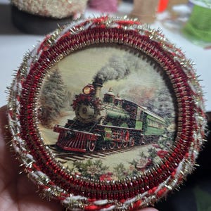 May include: Round Christmas ornament featuring a vintage train scene. The image is framed with red, white, and gold decorative trim. The train is green and red, with a plume of smoke rising from the engine.