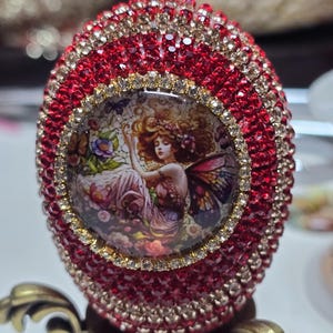 May include: An ornate, red jeweled egg with a central image of a fairy. The egg is covered in red and gold-toned crystals, with a gold-toned rim around the fairy image. The egg sits on a decorative, bronze-colored stand.