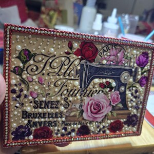 May include: Rectangular decorative box with a vintage sewing machine illustration, roses, and pearls. The box has a red and silver jeweled border and features text including "T. Ple Fournier" and "Bruxelles, Anvers".