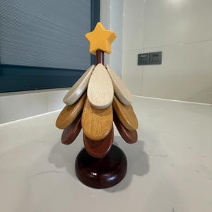 May include: A wooden Christmas tree decoration with a yellow star on top. The tree is made of wooden teardrop-shaped pieces in various shades of brown and beige, attached to a dark brown stem and base. The tree stands on a white surface.