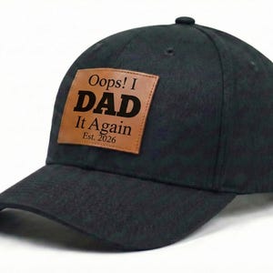 May include: A black baseball cap with a curved brim. A brown leather patch on the front displays the text "Oops! I DAD It Again Est. 2026" in bold, black letters. The cap has a button on top.