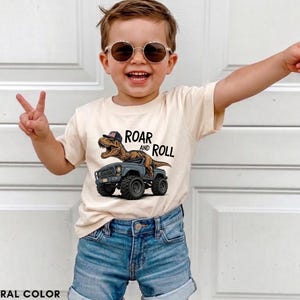 Puede incluir: Una camiseta color crema con un dinosaurio conduciendo un cami&oacute;n gris, con el texto "ROAR and ROLL". La camiseta se combina con pantalones cortos de mezclilla azules. La imagen muestra a un ni&ntilde;o peque&ntilde;o con gafas de sol haciendo el signo de la paz.