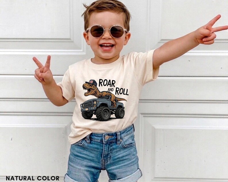Puede incluir: Una camiseta color crema con un dinosaurio conduciendo un cami&oacute;n gris, con el texto "ROAR and ROLL". La camiseta se combina con pantalones cortos de mezclilla azules. La imagen muestra a un ni&ntilde;o peque&ntilde;o con gafas de sol haciendo el signo de la paz.