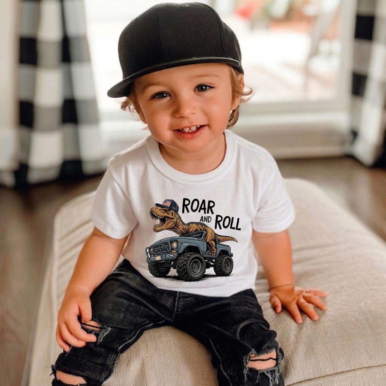 Puede incluir: Un ni&ntilde;o sonriente con una gorra negra y una camiseta blanca con un gr&aacute;fico de dinosaurio. La camiseta presenta un T-Rex de dibujos animados con una gorra conduciendo un cami&oacute;n azul, con las palabras "ROAR and ROLL". El ni&ntilde;o lleva vaqueros negros rotos.