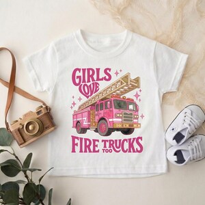 May include: White t-shirt with a pink fire truck graphic and the text "GIRLS LOVE FIRE TRUCKS TOO". The fire truck has a ladder and star accents. A toy camera and baby shoes are also in the image.