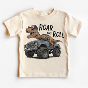 Puede incluir: Camiseta de color crema con un dinosaurio de dibujos animados que lleva una gorra con un dise&ntilde;o de bandera americana, conduciendo un cami&oacute;n monstruo azul. El texto "ROAR and ROLL" est&aacute; impreso encima del dinosaurio.