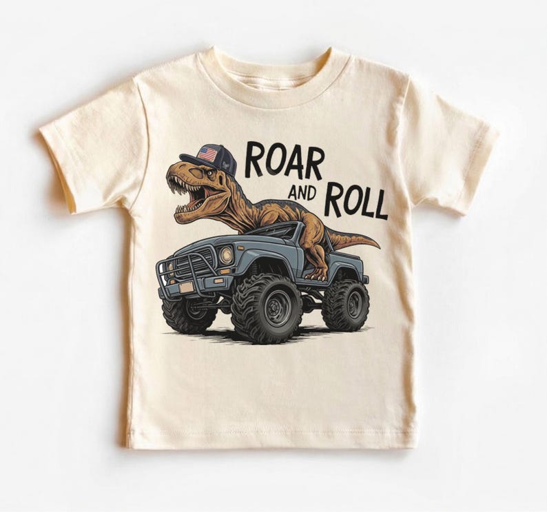 Puede incluir: Camiseta de color crema con un dinosaurio de dibujos animados que lleva una gorra con un dise&ntilde;o de bandera americana, conduciendo un cami&oacute;n monstruo azul. El texto "ROAR and ROLL" est&aacute; impreso encima del dinosaurio.
