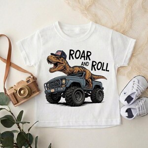 Puede incluir: Camiseta blanca de manga corta con un dinosaurio de dibujos animados que lleva una gorra y conduce un cami&oacute;n monstruo azul. El texto "ROAR and ROLL" est&aacute; impreso encima del cami&oacute;n. Tambi&eacute;n se ven unas zapatillas blancas y una c&aacute;mara de juguete.