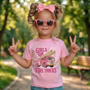 May include: A young child wearing a pink t-shirt with the text "GIRLS LOVE FIRE TRUCKS too" and a graphic of a pink fire truck. The child is also wearing pink sunglasses and a pink bow.