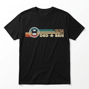 May include: A black short-sleeved t-shirt featuring a graphic of a floppy disk inside a circle, set against horizontal stripes in shades of orange, yellow, teal, and blue. The text reads: I KEEP ALL MY DAD JOKES IN A DAD-A-BASE.